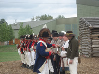 Photo:  Reenactors of “Captain Lewis Company” standing formation for their daily one gill of “rum” issue by the “orderly sergeant.” The two “civilians” (closest to the camera) represent the War Department contract employees who accompanied the “permanent party”: the hunter/ scout/ interpreter George Drouillard, and interpreter Toussaint Charbonneau (although the latter was never actually at River Du Bois), Sacagawea’s husband. 