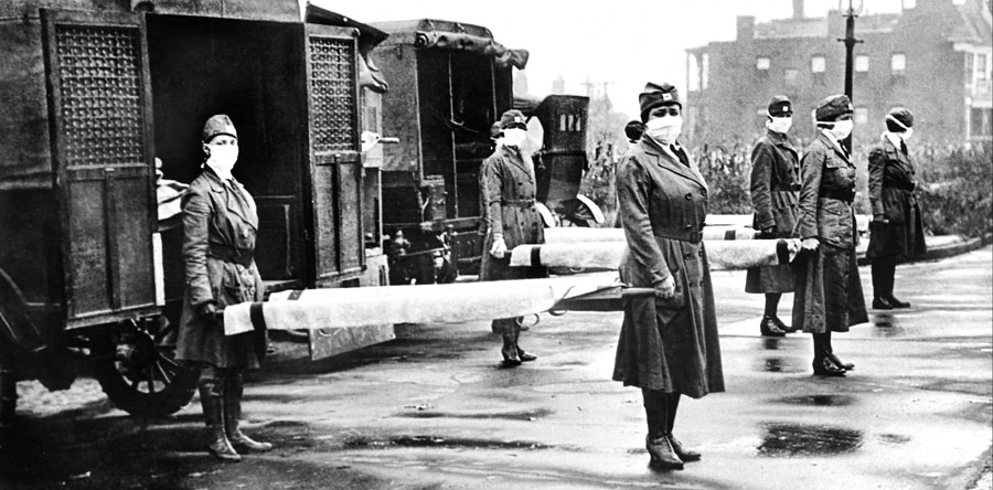 The image shows a group of St. Louis Red Cross Motor Corps workers on duty during the influenza epidemic in October 1918. The workers, all women, are dressed in military-style uniforms and are wearing face masks. They are standing in front of ambulances, each holding a stretcher, indicating their readiness to transport patients or supplies. The setting appears to be an urban area, with buildings visible in the background. The image is in black and white, and the atmosphere reflects the serious and urgent nature of their work during the epidemic. **Courtesy of the Library of Congress**.