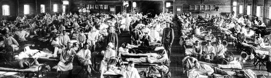 The image depicts a crowded emergency hospital at Camp Funston, Kansas, during the influenza epidemic in 1918. The large room is filled with rows of cots, each occupied by a soldier who appears to be ill. Medical personnel, including nurses and doctors, are seen attending to the patients. The room is packed with people, illustrating the scale and severity of the epidemic. The soldiers are dressed in hospital gowns, and some are wearing face masks. The setting conveys a sense of urgency and the overwhelming impact of the influenza outbreak on military personnel during this period. An emergency hospital at Camp Funston during the influenza epidemic, Kansas, 1918.