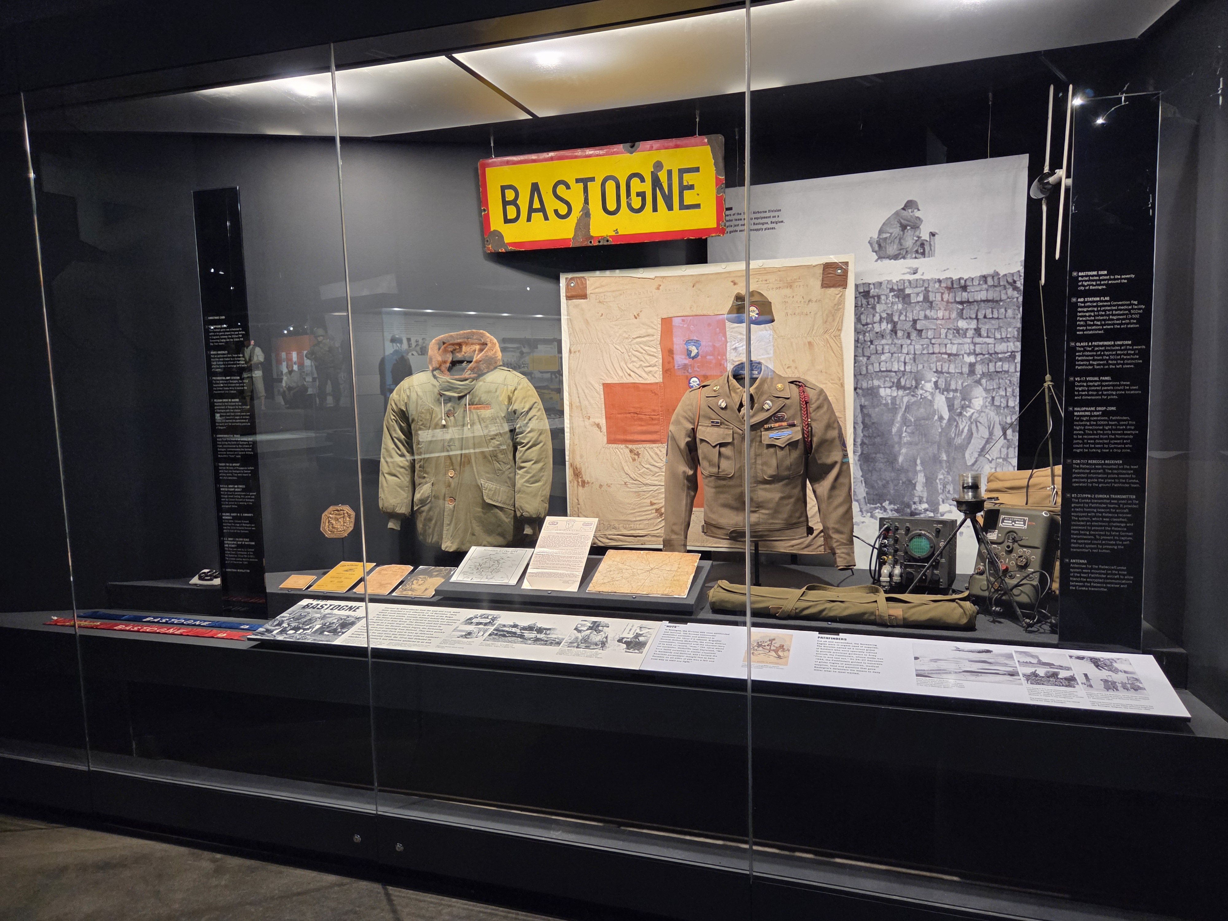 Museum exhibit featuring artifacts and uniforms from the Battle of the Bulge beneath a Bastogne road sign.